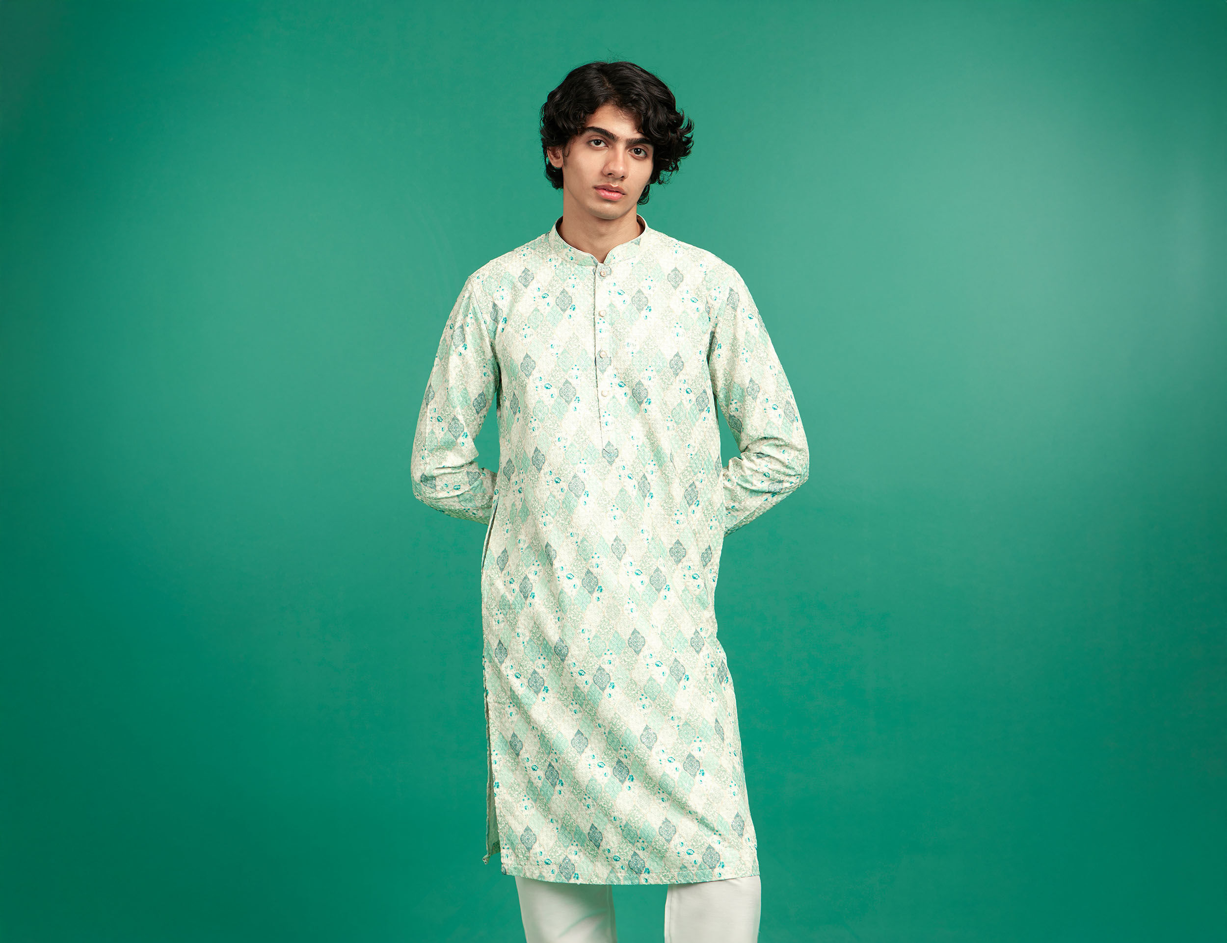 Young Indian man wearing a light green printed kurta with white churidar, posing against a solid green background